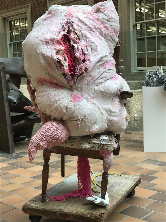 Sculpture made of fabril and crochet that looks like a muscle and skin mass that sits on a chair.