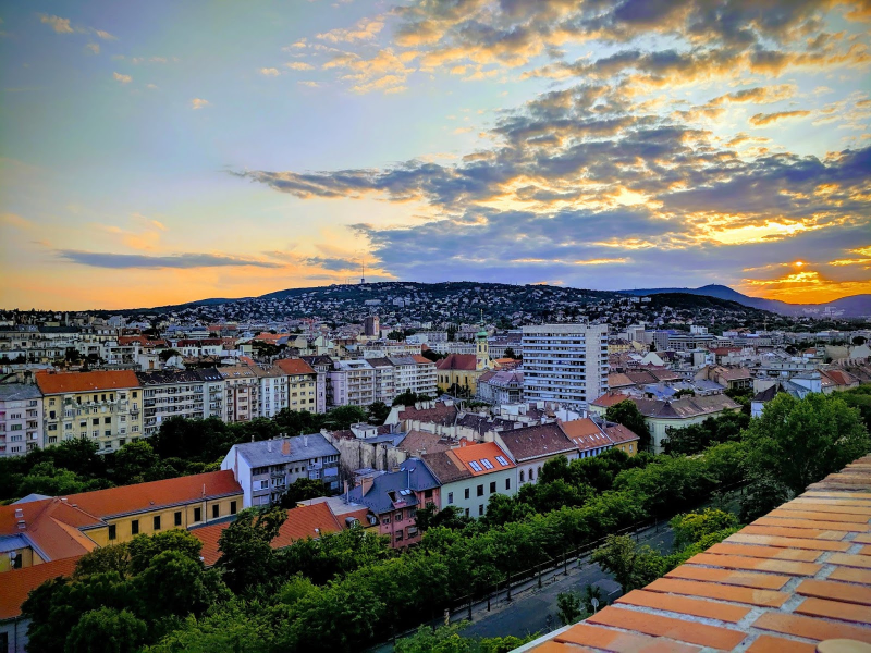 Sunsets in a semi cloudy sky behind a view of the Buda Hills. Building are in the forefront of the Buda Hills.