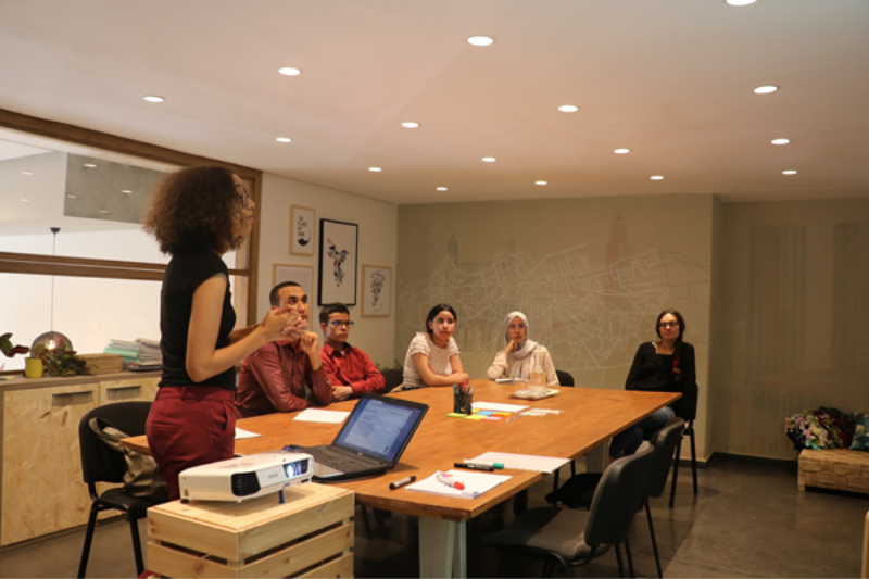 A presenter is standing in front of a table where five people are seated watching the presentation.