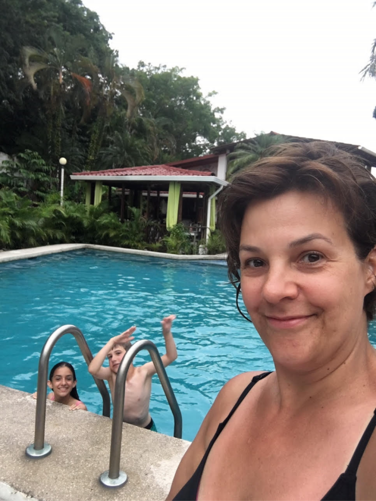 Debbie takes a selfie poolside with her children in the pool behind her.