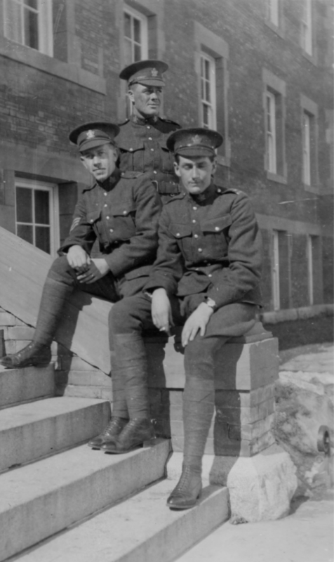 Two soldiers sit casually on the steps outside Athabasca Hall. Another soldier stands behind them looking into the distance.