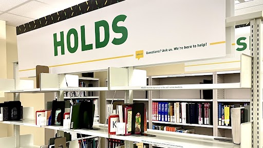 The holds section on the first floor of Cameron Library; designated by a giant sign above the bookshelf that reads 'Holds'.