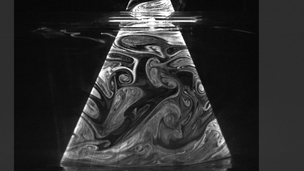 Grayscale visualization of turbulent airflow patterns using smoke structures within a sewer pipe cross-section.